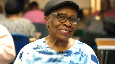 A woman wearing a blue and white patterned tie-dye top and a tweed flat cap, smiling at the camera. She has brown rimmed spectacles, and gold hoop earrings.