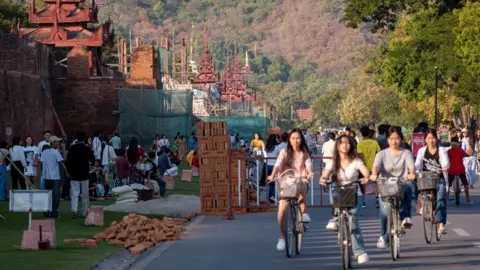 Jonathan Head/BBC Jovens mulheres andando de bicicleta em uma estrada que passa pelo palácio real em Mandalay. 