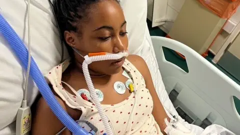 Family handout Daisy lies in a hospital bed and is hooked up to lots of tubes and devices. A white corrugated tube goes into her nose and she has several round patches on her chest with wires connected. She wears a cream vest with hearts on and her hair is scraped back into a pony tail.
