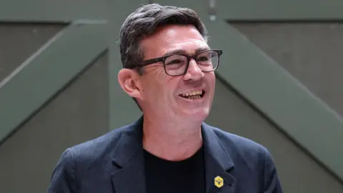 Andy Burnham is smiling and looking slightly off to the left. His short hair is dark brown, with some greys coming through. He's wearing dark rimmed glasses and a dark blue blazer with a black t-shirt underneath. 