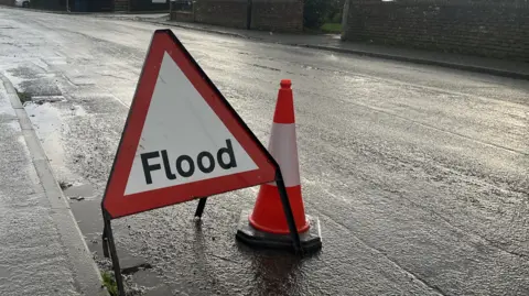 A red and white triangular road sign on a road, next to a red and white cone. The road is wet. The sign reads "flood".
