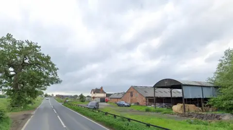 A long, straight, road with a tree at one side and agricultural buildings on the other side.