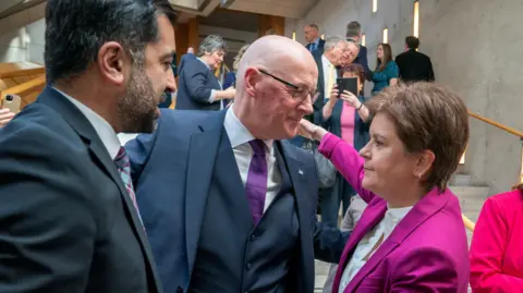 PA Media In the grey and light wood lobby of the Scottish Parliament, Humza Yousaf, John Swinney and Nicola Sturgeon are standing talking. Swinney is warmly embracing Sturgeon, who is staring beyond him. Yousaf stands off to the side. There is a crowd of people on the stairs behind them. Someone is taking a picture.