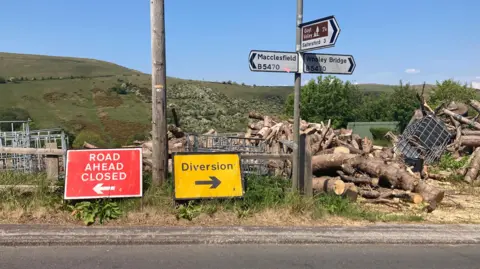 A view of the road between Macclesfield and Whaley Bridge. It has signs saying the road is closed ahead and diversion route, in front of the signs pointing the way to both towns