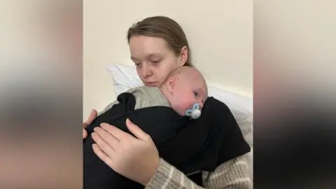 A woman looks ill while hugging her newborn baby. Both are covered in blankets. The baby has a dummy in its mouth.