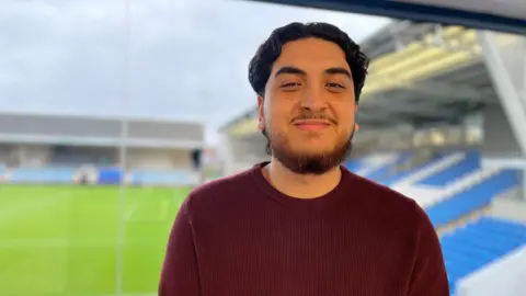 Emma Baugh/BBC A head-and-shoulders picture of Joseph Lucak. He has medium-length black hair and a beard and is smiling at the camera. He is wearing a dark jumper. Terracing with blue seats and the green pitch can be seen through a window behind him.