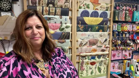 James Grant/BBC Woman wearing pink and black top stands in front of shelves of wrapping paper.