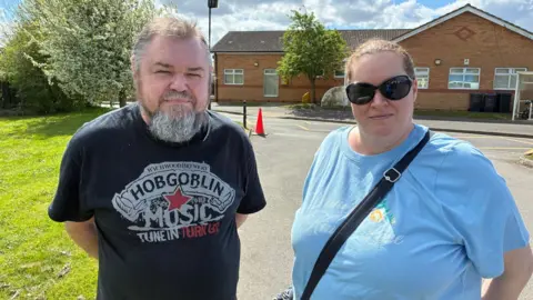 Paul wears a Hobgoblin music black tshirt and has a goatee beard.  Jo wears sunglassess and a blue top with a cross-body bag.