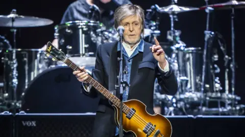 PA Paul McCartney performing on stage recently. He wears a black suit with a blue shirt underneath and holds a brown guitar as he stands in front of a microphone.