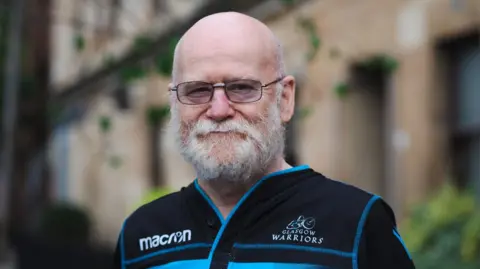 Person wearing a black Glasgow Warriors rugby shirt with blue stripes and Macron logo, standing outdoors in front of a stone building with greenery