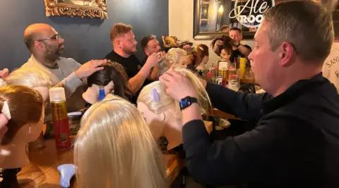 Pub scene with dads sat around a table. In front of them are mannequin heads with hair. The men are being taught how to do plaits. 