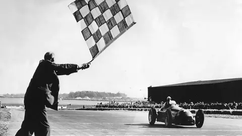 Getty Images A black and white photo of the 1950 British Grand Prix at Silverstone. An Alpha Romeo with a thin bullet-shaped body, with a driver's head visible above the windscreen, passes the finish line as a man in a dark-coloured coat waves a chequered flag. Spectators are visible behind a low barrier at the edge of the track, with a stand behind them.