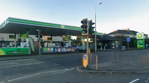 Shell petrol station on roadside. It is mostly green and there are a set of traffic lights in the foreground