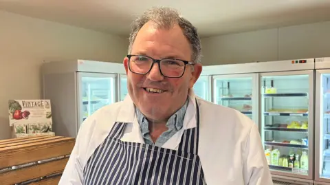 BBC Peter Themans looks into the camera and smiles. He has short dark grey hair and is wearing a pair of black-framed glasses. He is wearing a light blue collared shirt, a long white coat over it, and a navy blue and white striped butcher's apron. He is stood inside the shop, in front of five glass-fronted large fridges with produce in them. The walls of the shop are painted cream. 