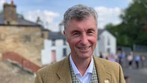 Niall Hammond with grey hair, wearing a mustard tweed blazer and white shirt, smiles at the camera.