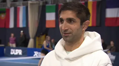 James Chaudry giving an interview. He is a wearing a white hoody. He is looking away from the camera. The flags of various European countries are seen hanging in a wall behind him. One player could also be seen on the court.