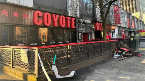 BBC A nightclub with the words COYOTE in red and the letters GLY seen on the far right. Broken bits of charred wood and red lettering from the sign lay on the floor outside. 