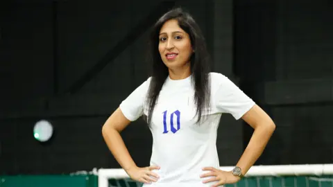 Sana Gill Sana, an Asian woman, wearing a white jersey with the number "10" written in blue in the middle. She has her hands on her hips, with a watch on her left wrist, and is looking at the camera with a smile. The background is dark.