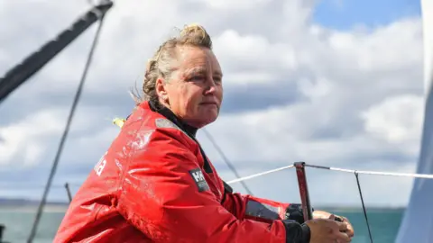 PA Media Pip Hare looks over the edge of her yacht toward the sea as she fastens a rope around a metal pole. Her blonde and grey hair is windswept and she is wearing a large red waterproof coat