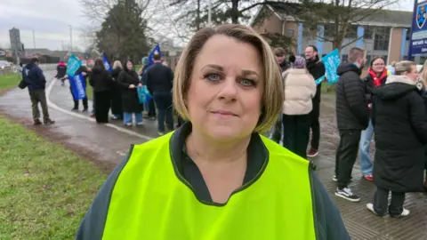 A woman in her 40s with a short dirty blonde bob. She is wearing makeup, a navy fleece, and a green hi-vis tabard as she stands in front of a picket line of teachers and staff outside a school. The teachers are wrapped up warm with coats and hats in the cold weather, and are holding up blue union flags.