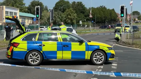 A police car with its boot open is parked across a road behind blue and white police tape. Police officers are talking to each other and inspecting the scene, by a pedestrian crossing with traffic lights. Other vehicles can be seen parked in the background.