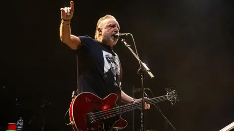  Peter Hook playing a red bass and singing into a microphone- he wears a black T-shirt with Oasis on the front - he's performing at Wembley arena