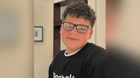 Family photograph Carter with brown hair wearing black glasses wearing a black jumper and white collar. He is in a living room with a bookcase behind him and a framed picture on the wall. He is smiling.