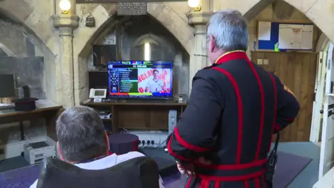 Two men watch a television showing an England football screen inside a stone-walled room with arched ceilings and wooden furniture.