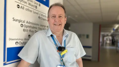 Jessica Lane/BBC Merv Denton wearing his work uniform - a light blue shirt and dark blue NHS lanyard - and standing in a hospital corridor. He has short brown hair and is leaning against the wall and smiling.