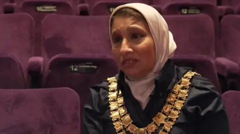 BBC Solihull's mayor, Councillor Shahin Ashraf, is pictured sitting in a theatre with purple velvet seats to the side and behind her. She is wearing a light pink headscarf and two large gold chains over a black jacket.