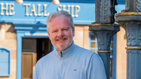 BBC Studios Drama A person stands outdoors beside a decorative cast‑iron column, wearing a light blue sweater and jeans.
Behind them is a stone building with bright blue trim and a sign reading “The Tall Ship” above the doorway.