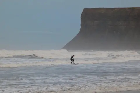 BBC Weather Watchers / Helen Earth A surfer, in a full body-suit, rides a wave in front of a cliff.