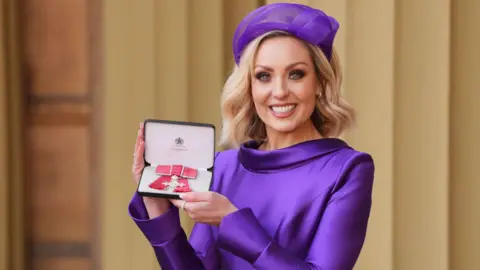 Amy Dowden smiles as she holds her MBE in its presentation box. She is wearing a purple silk dress with a matching purple hat.