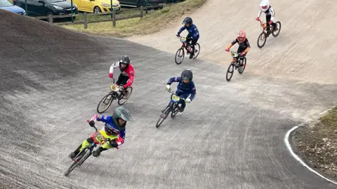 Ben Fisher Archie is leading a group of six racers on a banked turn. He is wearing a blue top with fluorescent yellow leggings and has a grey helmet on his head.