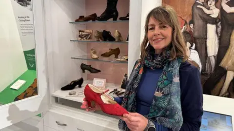 A woman with brown hair, a blue top and a blue and green scarf is holding a red high heel from the 1920s. there is a shelf of high heels in the background and a painting on the wall