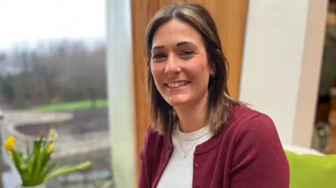 Head and shoulders image of Kirsty Speers. She has shoulder-length, brown hair and is smiling at the camera. She is wearing a white top with a burgundy cardigan. Beside her is a vase containing yellow flowers. 