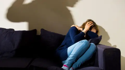 A woman wearing a navy jumper and light blue denim skinny jeans cowers in fear on a navy sofa as the shadow of a man with his fist raised looms over her.