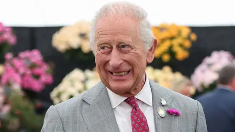 PA Media Head and shoulders of a smiling King Charles who is wearing a pale grey suit, white shirt and burgundy patterned tie. He is looking to the left and standing in front of bouquets of flowers