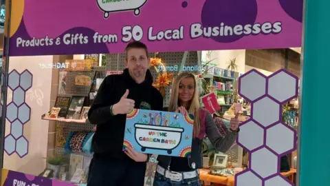 Dave and Kez are stood in front of the Unity shop window holding a "Created by Up the Garden Bath" sign in their hands. Dave is wearing a black hoodie with the Garden Bath logo on the front right and is giving a thumbs up to the camera. Kez, who has long blonde hair,  is doing the same thumbs up gesture. Behind them in the shop window a sign reads "Products and gifts from 50 local businesses". 