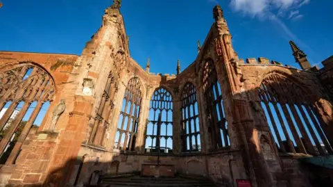 Getty Images The ruins of Coventry's old cathedral include a curved structure and tall arched windows. The ruins are seen against a blue sky.