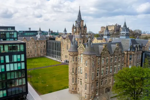 Getty Images A four storey sandstone building with a grass lawn in front of it adjacent to a tall modern tower made of steel and glass