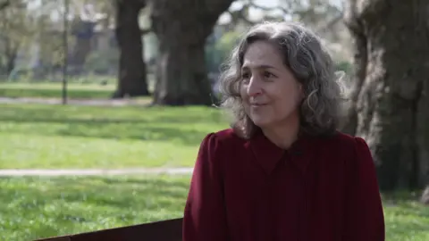 Caroline Woodley sitting in a park with grass and tree trunks behind her. She is wearing a dark red top