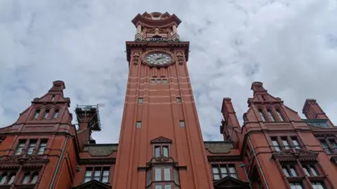 Image shows the red-brick facade of the Kimpton Clocktower hotel, with the clocktower itself rising up in the centre of the frame. 