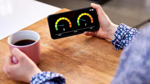 BBC A woman wearing a floral blouse and jumper holds a mobile phone showing a reading from their smart meter in one hand and a cup of black coffee in a pink mug in the other. Her arms are resting on a wooden table top.
