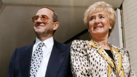 A man and woman stand outside a building entrance beneath a decorative canopy. Both are dressed in formal attire, the man in a dark suit and brown glasses and the woman in a patterned jacket with gold trim. The photo is old.