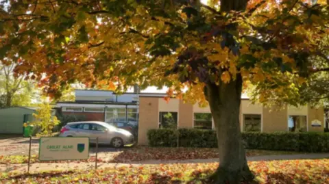 A large tree with red, yellow and orange autumn leaves on it and leaves on the ground in front of a beige, one-storey building with a silver car parked alongside