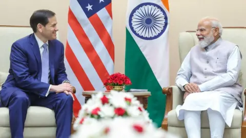 Marco Rubio (left) and Narendra Modi (right) smile at each other as they sit beside their respective countries' flags.