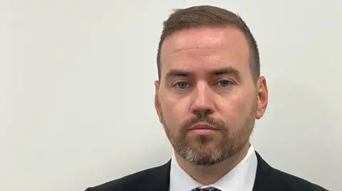 A head and shoulders picture of Basildon Council leader Gavin Callaghan. He is looking at the camera and wearing a black suit jacket, a white shirt and a black and white tie.