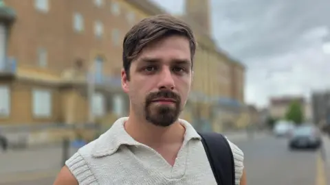Paul Moseley/BBC Alex Catt is wearing a short-sleeve, cream top. We can see a black bag strap over his left shoulder. Behind him is Norwich City Hall.