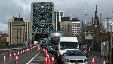 A lane of traffic crosses the Tyne Bridge. There are red traffic cones on the left side of the bridge, blocking one lane.